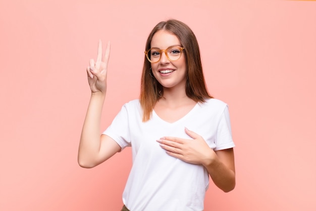 Junge rothaarige Frau, die glücklich, zuversichtlich und vertrauenswürdig aussieht, lächelt und Siegeszeichen zeigt, mit einer positiven Haltung gegen flache Wand