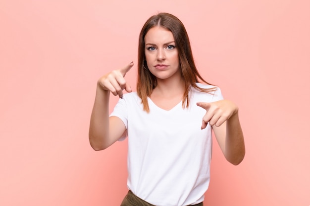 Junge rothaarige Frau, die mit beiden Fingern und wütendem Ausdruck nach vorne zeigt und Ihnen sagt, Sie sollen Ihre Pflicht gegen flache Wand tun