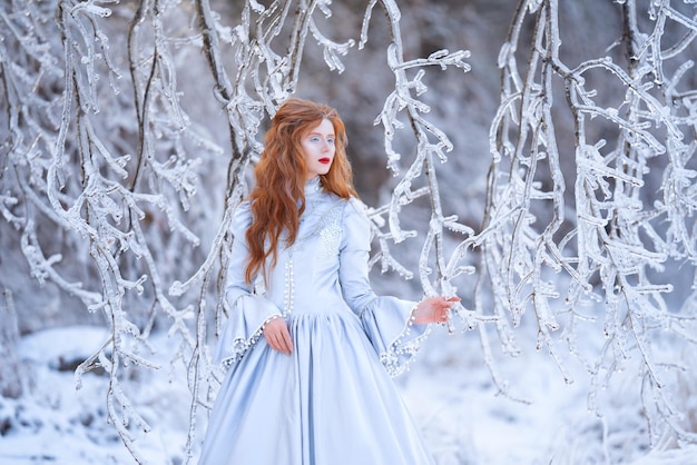 Junge rothaarige Frau, eine Prinzessin, geht in einem blauen Kleid in einem Winterwald spazieren