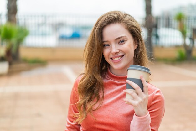 Junge rothaarige Frau im Freien hält mit glücklichem Gesichtsausdruck einen Kaffee zum Mitnehmen in der Hand