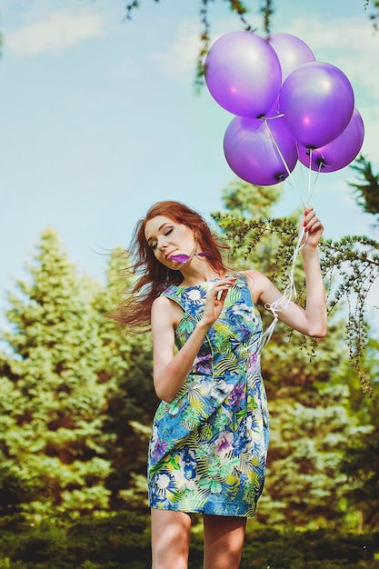 junge rothaarige Frau im Frühlingsgarten mit Sommersprossen. Lila Luftballons. Geburtstag, Feiertagsfeier.