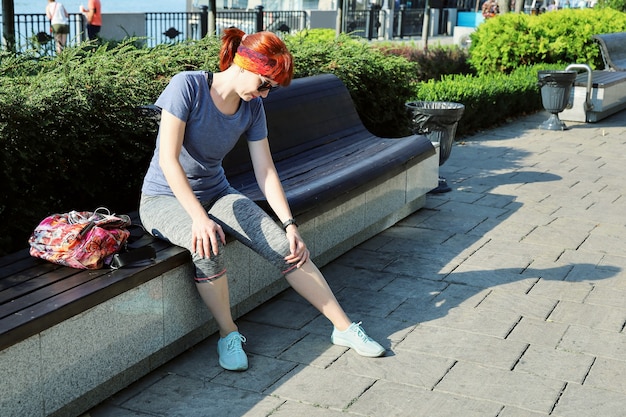 Junge rothaarige Frau in Sportkleidung sitzt draußen auf der Bank und berührt ihr schmerzendes Knie