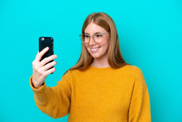 Junge rothaarige Frau isoliert auf blauem Hintergrund, die ein Selfie mit Handy macht