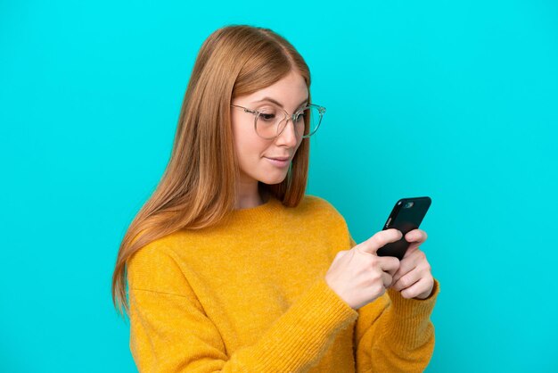 Junge rothaarige Frau isoliert auf blauem Hintergrund, die eine Nachricht oder E-Mail mit dem Handy sendet