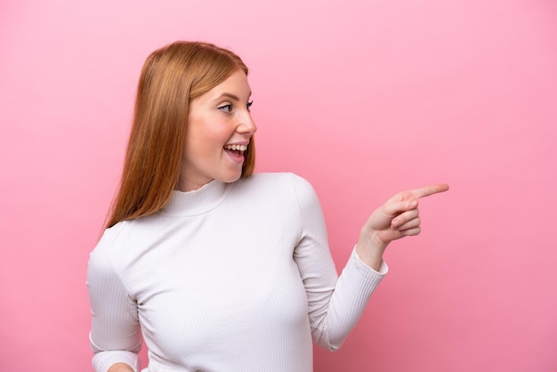 Junge rothaarige Frau isoliert auf rosa Hintergrund, die mit dem Finger zur Seite zeigt und ein Produkt präsentiert