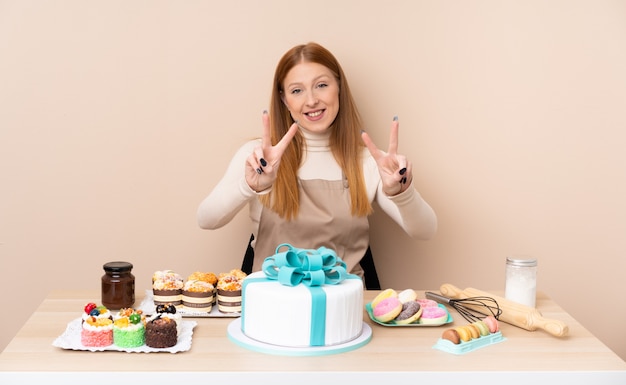 Junge rothaarige Frau mit einem großen Kuchen, der lächelt und Siegeszeichen zeigt