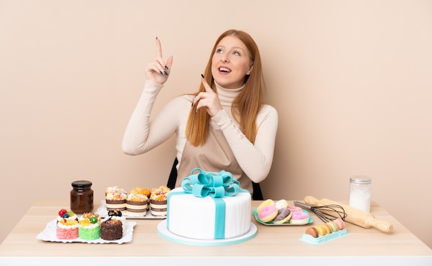 Junge rothaarige Frau mit einem großen Kuchen, der mit dem Zeigefinger eine große Idee zeigt