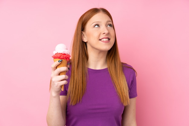 Junge rothaarige Frau mit einem Kornett-Eis über der isolierten rosa Wand, die beim Lächeln nach oben schaut