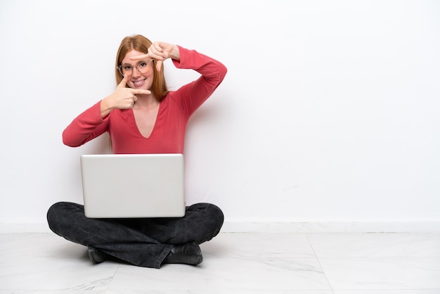 Junge rothaarige Frau mit einem Laptop, der auf dem Boden sitzt, isoliert auf weißem Hintergrund, der das Gesicht fokussiert Framing-Symbol