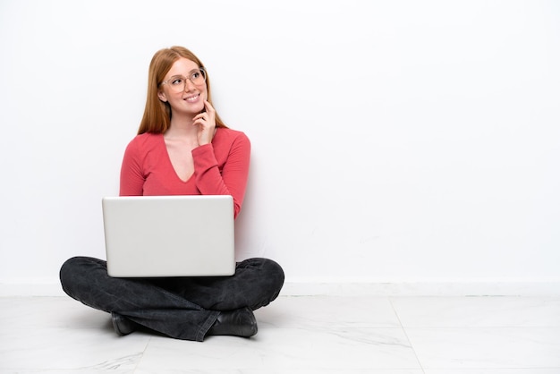 Junge rothaarige Frau mit einem Laptop, der isoliert auf weißem Hintergrund auf dem Boden sitzt und eine Idee denkt, während sie nach oben schaut