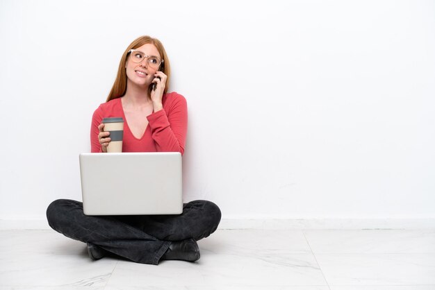 Junge rothaarige Frau mit einem Laptop, der isoliert auf weißem Hintergrund auf dem Boden sitzt und Kaffee zum Mitnehmen und ein Handy hält