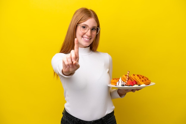 Junge rothaarige Frau mit Waffeln isoliert auf gelbem Hintergrund, die einen Finger zeigt und hebt