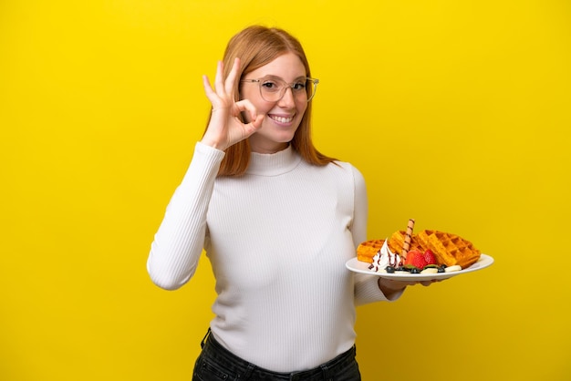 Junge rothaarige Frau mit Waffeln isoliert auf gelbem Hintergrund, die mit den Fingern ein Ok-Zeichen zeigt