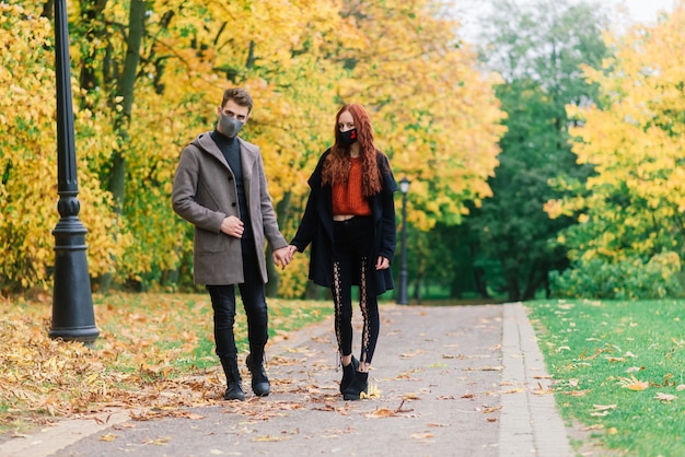 Junge rothaarige Frau setzt eine Gesichtsmaske auf, während sie mit jungem Mann im Herbstpark geht.