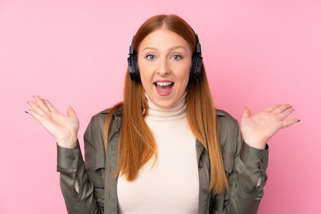 Junge rothaarige Frau über isolierter rosa Wand überrascht und Musik hören