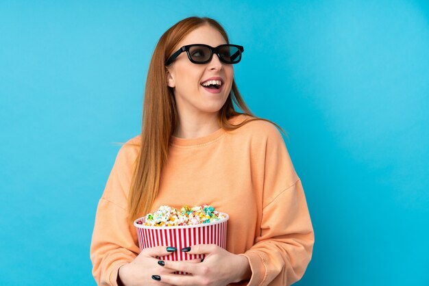 Junge rothaarige frau über isolierter wand mit 3d-brille und hält einen großen eimer popcorn