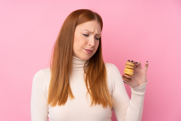 Foto junge rothaarige frau über rosa, die bunte französische macarons mit traurigem ausdruck hält