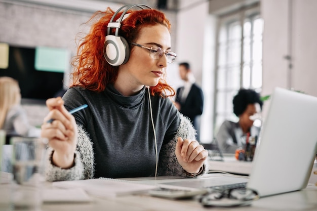 Junge rothaarige Geschäftsfrau, die Musik über Kopfhörer genießt, während sie bei der Arbeit eine E-Mail auf dem Computer liest, hier sind Menschen im Hintergrund