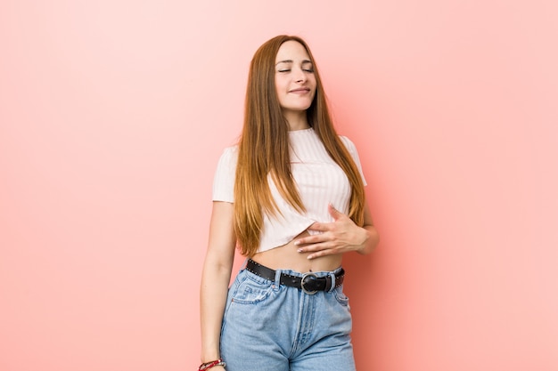 Junge rothaarige Ingwerfrau gegen eine rosa Wand berührt Bauch, lächelt sanft