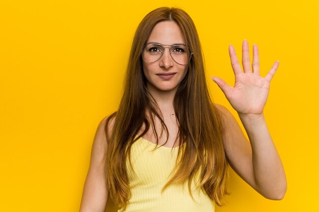 Junge rothaarige Ingwerfrau mit Sommersprossen, die fröhlich lächeln und Nummer fünf mit den Fingern zeigen.