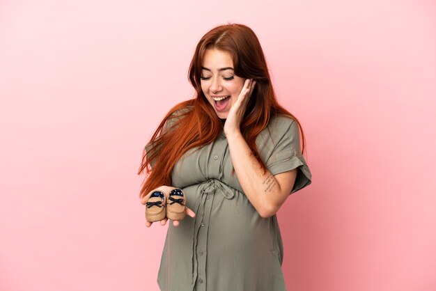 Junge rothaarige kaukasische Frau isoliert auf rosa Hintergrund schwanger und hält Babyschuhe mit überraschtem Ausdruck