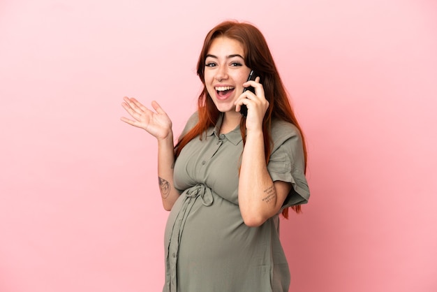 Junge rothaarige kaukasische Frau isoliert auf rosa Hintergrund schwanger und mit Handy mit überraschtem Ausdruck