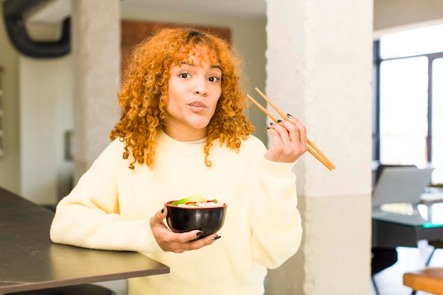 Junge rothaarige lateinische hübsche Frau, die zu Hause Ramen-Nudelschüssel isst