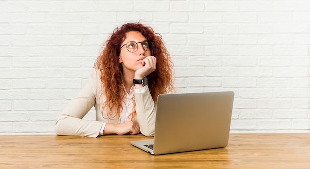 Junge rothaarige lockige Frau, die mit ihrem Laptop arbeitet, der mit zweifelhaftem und skeptischem Ausdruck seitwärts schaut.