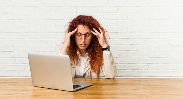 Junge rothaarige lockige Frau, die mit ihrem Laptop arbeitet, der Schläfen berührt und Kopfschmerzen hat.