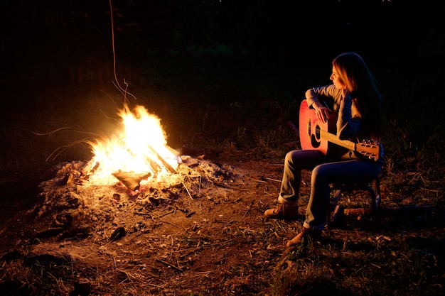 Junge rothaarigefrau, die gitarre nahe feuer am nachtlager spielt