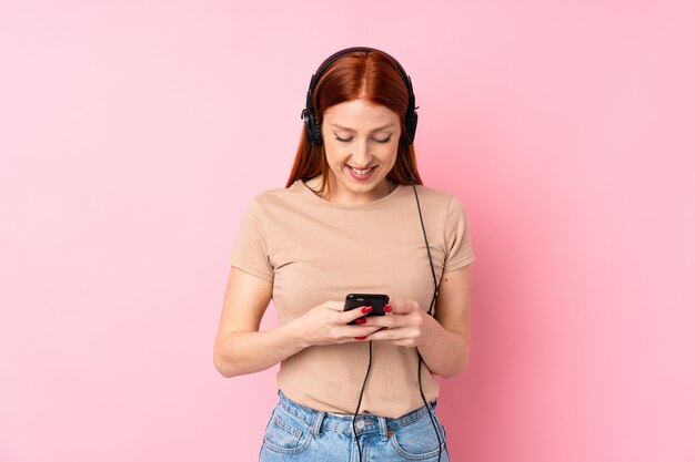 Junge Rothaarigefrau über lokalisierter rosa Wand unter Verwendung des Mobiles mit Kopfhörern
