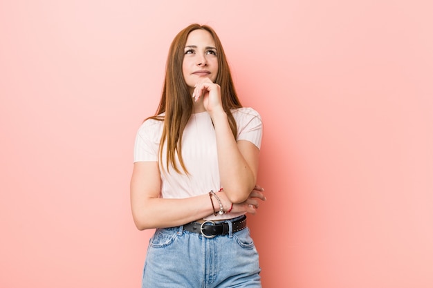 Junge Rothaarigeingwerfrau gegen eine rosa Wand, die seitlich mit zweifelhaftem und skeptischem Ausdruck schaut.