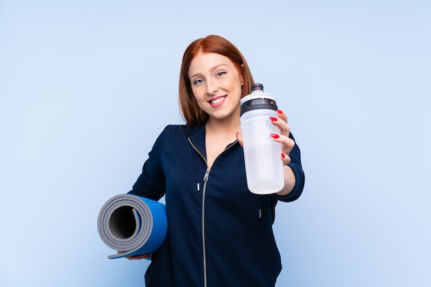 Junge Rothaarigesportfrau über lokalisiertem Blau mit Sportwasserflasche und mit einer Matte