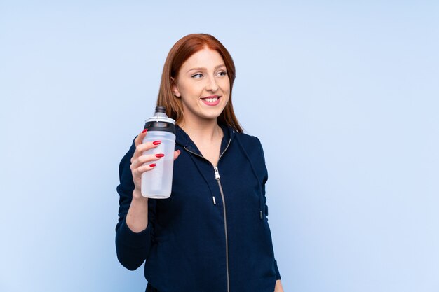 Junge Rothaarigesportfrau über lokalisierter blauer Wand mit Sportwasserflasche