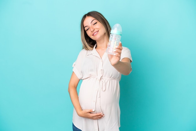 Junge rumänische Frau isoliert auf blauem Hintergrund schwanger und hält eine Babyflasche