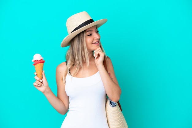 Junge rumänische Frau mit Eis und Strandtasche isoliert auf blauem Hintergrund, denkt an eine Idee und schaut zur Seite