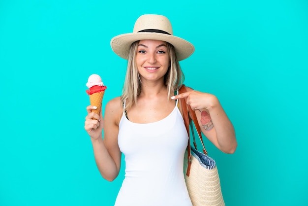 Junge rumänische Frau mit Eis und Strandtasche isoliert auf blauem Hintergrund mit überraschtem Gesichtsausdruck