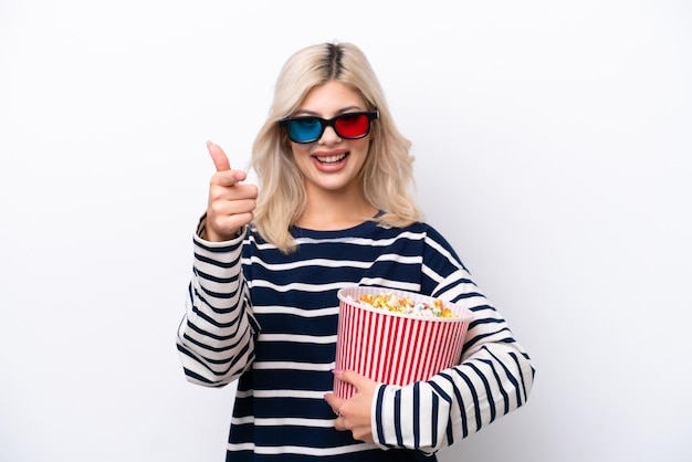 Junge Russin isoliert auf weißem Hintergrund mit 3D-Brille und hält einen großen Eimer Popcorn, während sie nach vorne zeigt