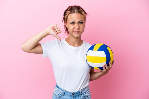 Junge Russin spielt Volleyball isoliert auf rosa Hintergrund und zeigt Daumen nach unten mit negativem Ausdruck