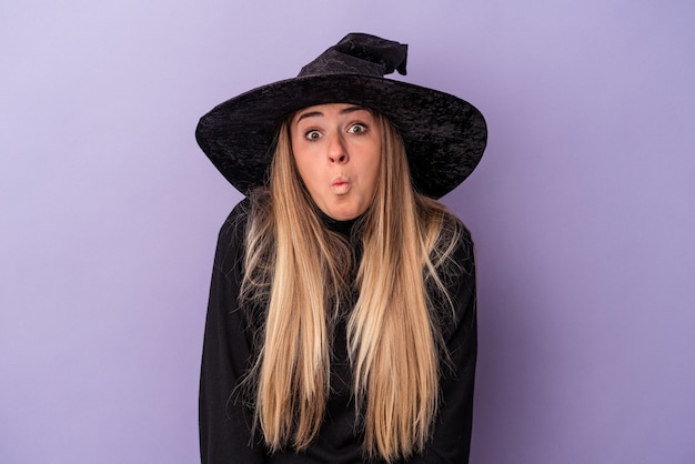 Junge russische Frau, die als Hexe verkleidet ist, die Halloween isoliert auf violettem Hintergrund feiert, zuckt mit den Schultern und öffnet die Augen verwirrt.