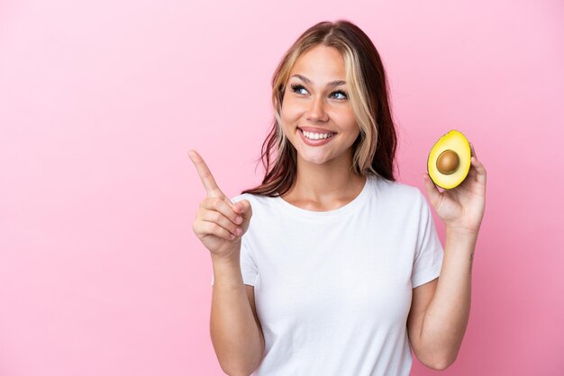 Junge russische Frau, die Avocado isoliert auf rosafarbenem Hintergrund hält und beabsichtigt, die Lösung zu realisieren, während sie einen Finger hochhebt