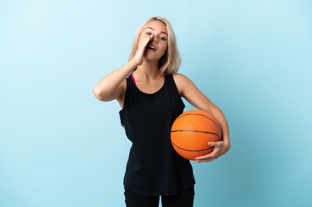 Junge russische Frau, die Basketball spielt, isoliert auf blauer Wand, die etwas schreit und ankündigt