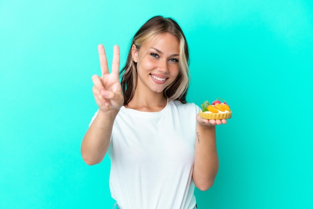 Junge russische Frau, die eine Fruchtbonbons lokalisiert auf blauem Hintergrund hält, lächelt und zeigt Victory-Zeichen