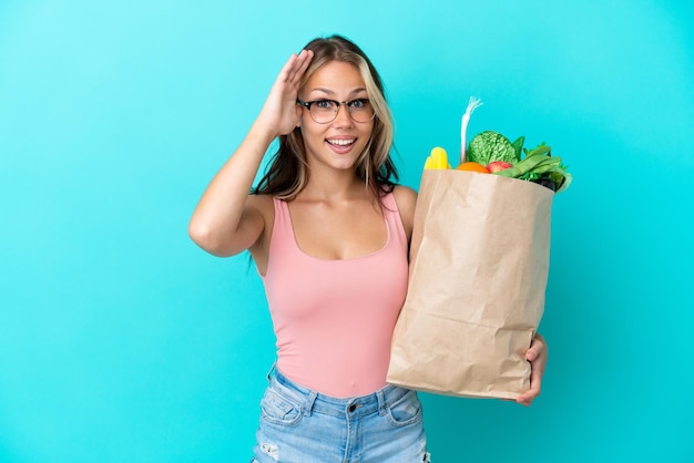 Junge russische Frau, die eine Lebensmitteleinkaufstasche isoliert auf blauem Hintergrund mit überraschtem Ausdruck hält