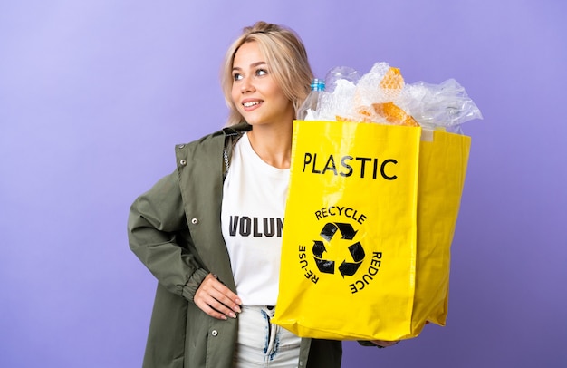 Junge russische Frau, die eine Recycling-Tasche voll Papier hält, um lokalisiert auf lila, das mit Armen an der Hüfte und lächelnd aufbereitet zu recyceln