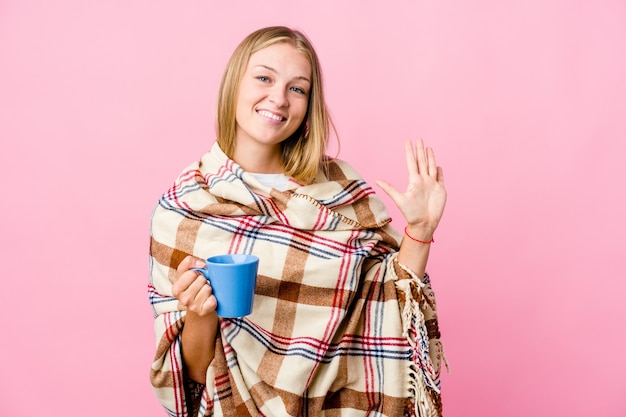 Junge russische Frau, die in eine Decke gewickelt wird, die Kaffee trinkt, der fröhlich lächelt und Nummer fünf mit den Fingern zeigt.