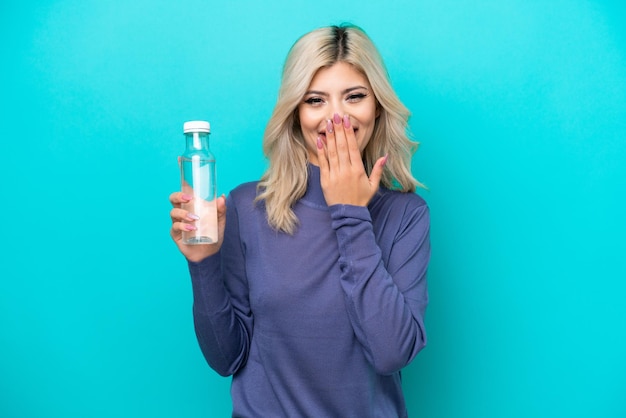 Junge russische Frau mit einer Flasche Wasser isoliert auf blauem Hintergrund glücklich und lächelnd, den Mund mit der Hand bedeckend