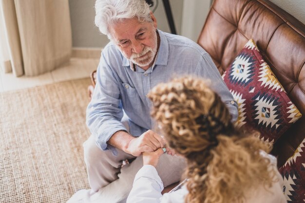 Junge Ärztin, die älteren Mann zu Hause überprüft und hilft. Arzt hilft älteren Patienten und kümmert sich, sitzt auf dem Sofa. Heimpflegerin mit pensioniertem alten Mann zu Hause