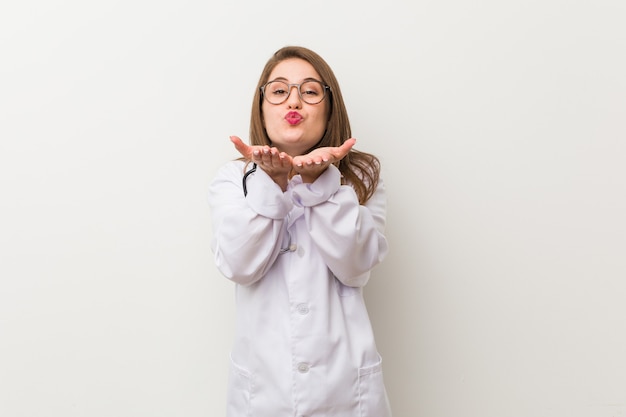 Junge Ärztin gegen eine weiße Wand, die Lippen faltet und Handflächen hält, um Luftkuss zu senden.