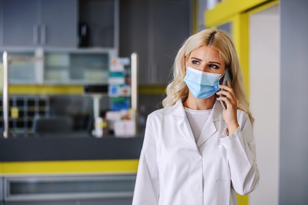 Junge Ärztin in steriler weißer Uniform, die im Krankenhaus steht und einen wichtigen Telefonanruf hat.